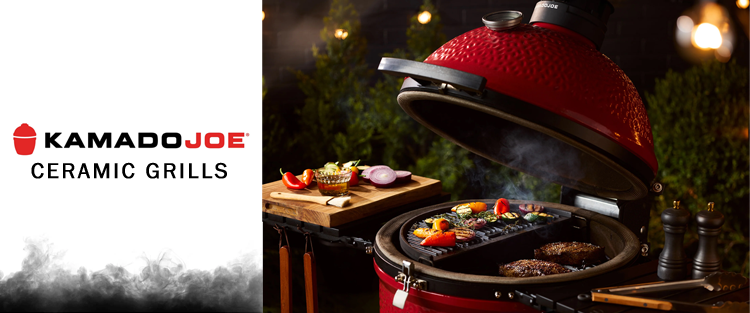 Red Kamado Joe ceramic grill with food on a wooden cutting board, surrounded by greenery.